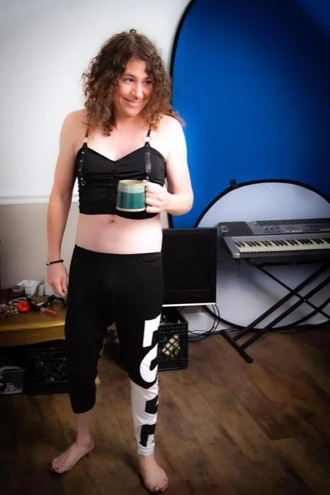 B, a woman with long curly brown hair, smiling and looking off to one side, holding a green mug in a room with a blue backdrop and a keyboard on a stand